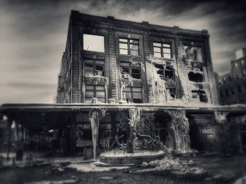 Abandoned industrial building with destroyed windows and an icicle-covered facade behind a deteriorated canopy, in black and white.