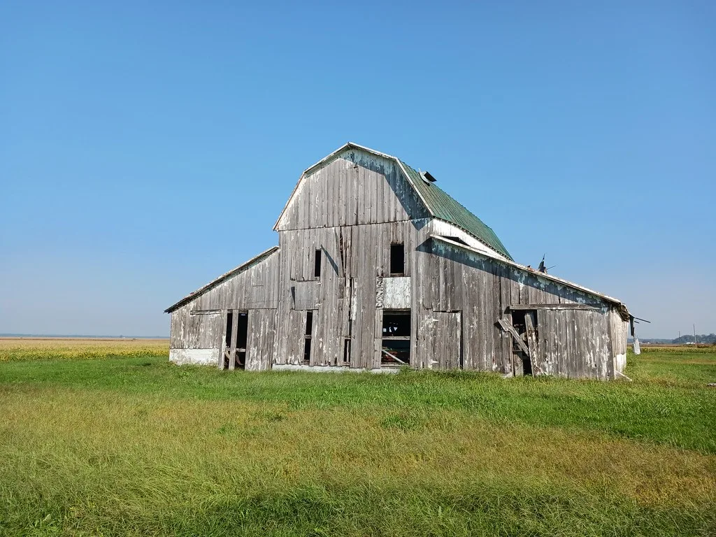 Large abandoned gray wooden barn with broken doors and a damaged green roof in a meadow.