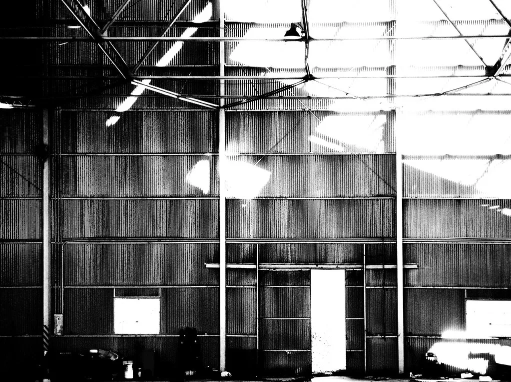Interior of a metal hall with holes in its roof, in black and white.
