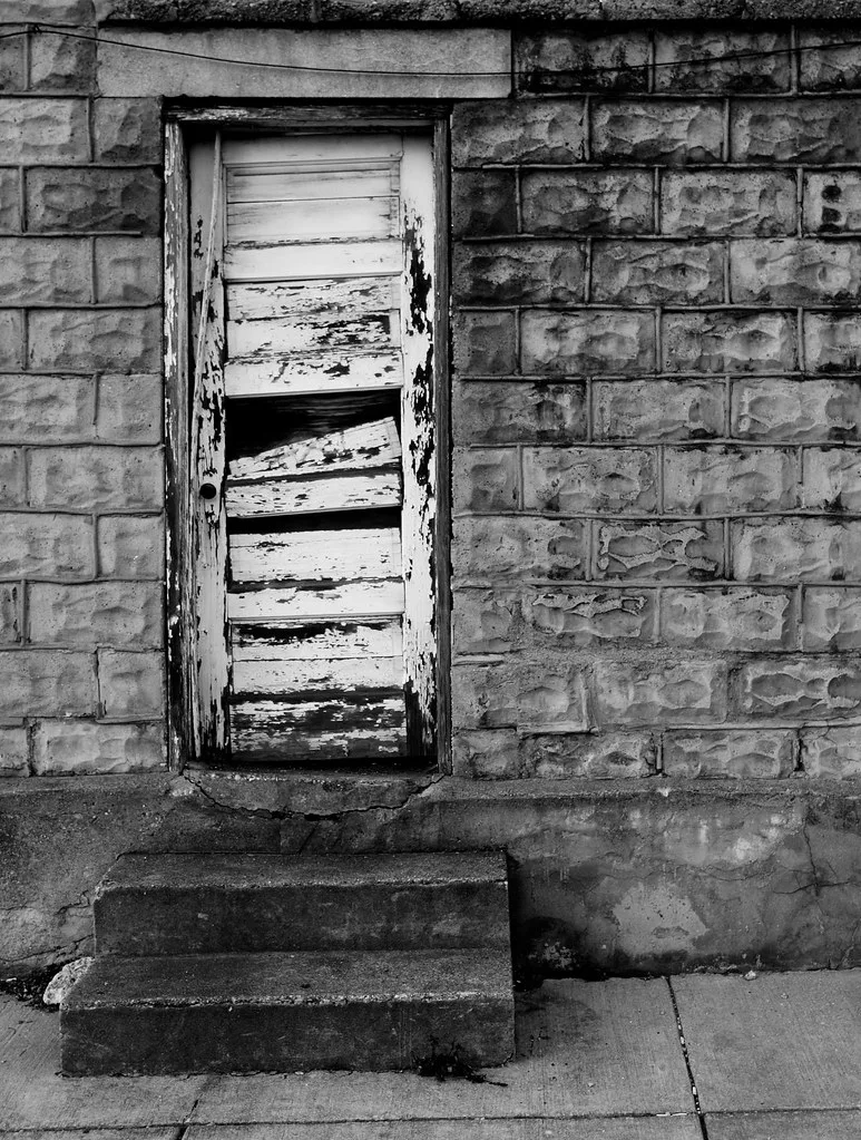 Derelict door with separated wooden boards above three concrete steps.