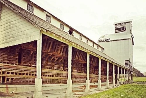 Long wooden industrial building open to its internal framing, with a tower at the far end.