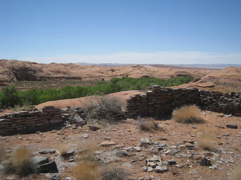 Ruined walls of flat stones above a desert valley and a ribbon of green trees.