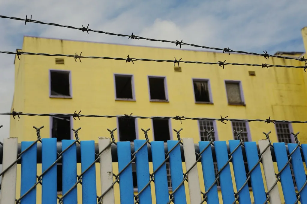 Yellow building with broken windows behind a barbed-wire fence.