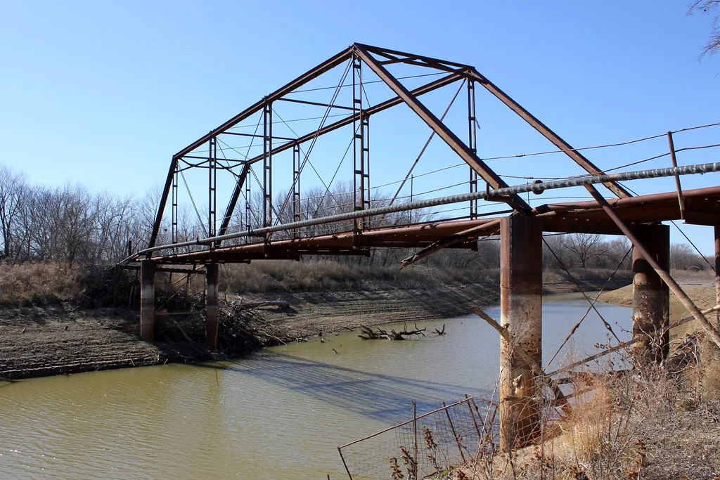 Ancien pont abandonné à treillis rouillé sans tablier au-dessus d’une rivière boueuse.
