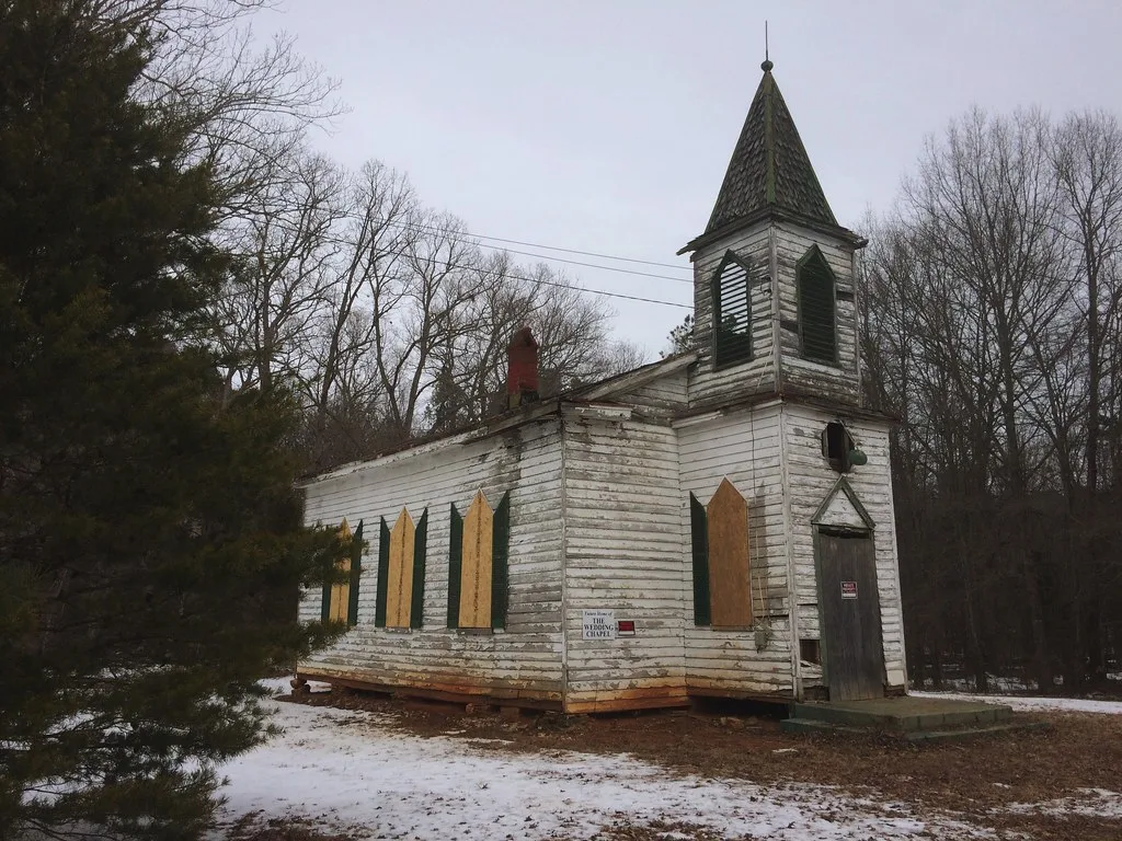 Small white church with a pointed bell tower and boarded windows on partly snow-covered ground.