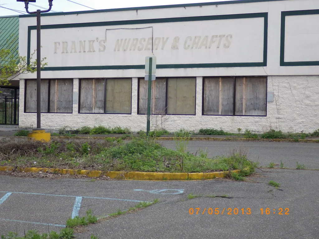Ancien magasin aux vitrines condamnées devant un parking envahi d’herbes.
