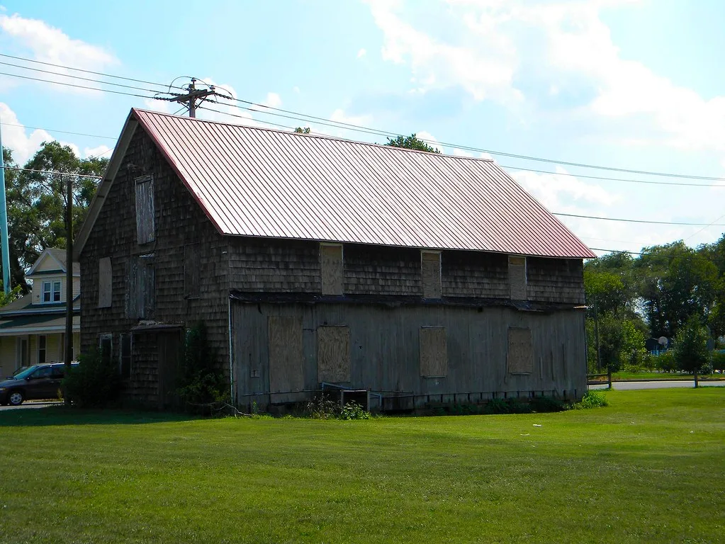 Large dark wooden building with boarded windows beneath a red metal roof.