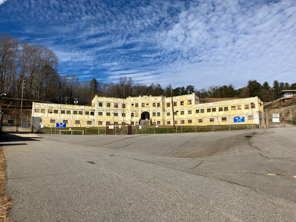 Old yellow prison with boarded windows behind a fenced enclosure.