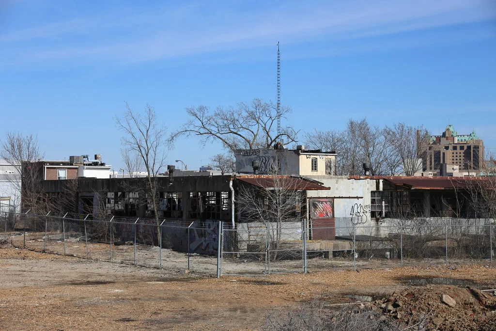 Abandoned former industrial building with broken window bays and graffiti-covered walls behind a wire fence.