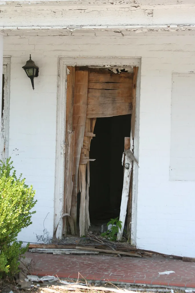 Entrance of a white house with a shattered wooden door and fallen boards on the porch.