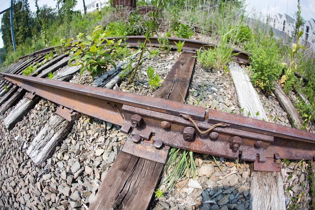 Close view of an abandoned railway track with rusty rails and split sleepers overgrown with plants in the ballast.