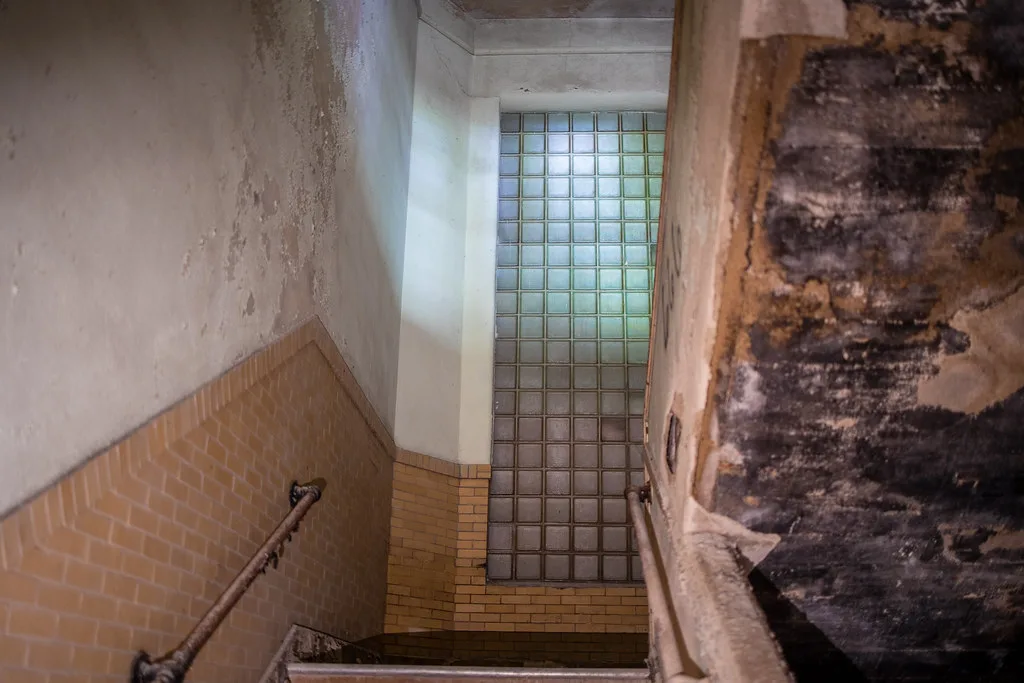 Old stairway with damaged walls facing a tall glass-block window.