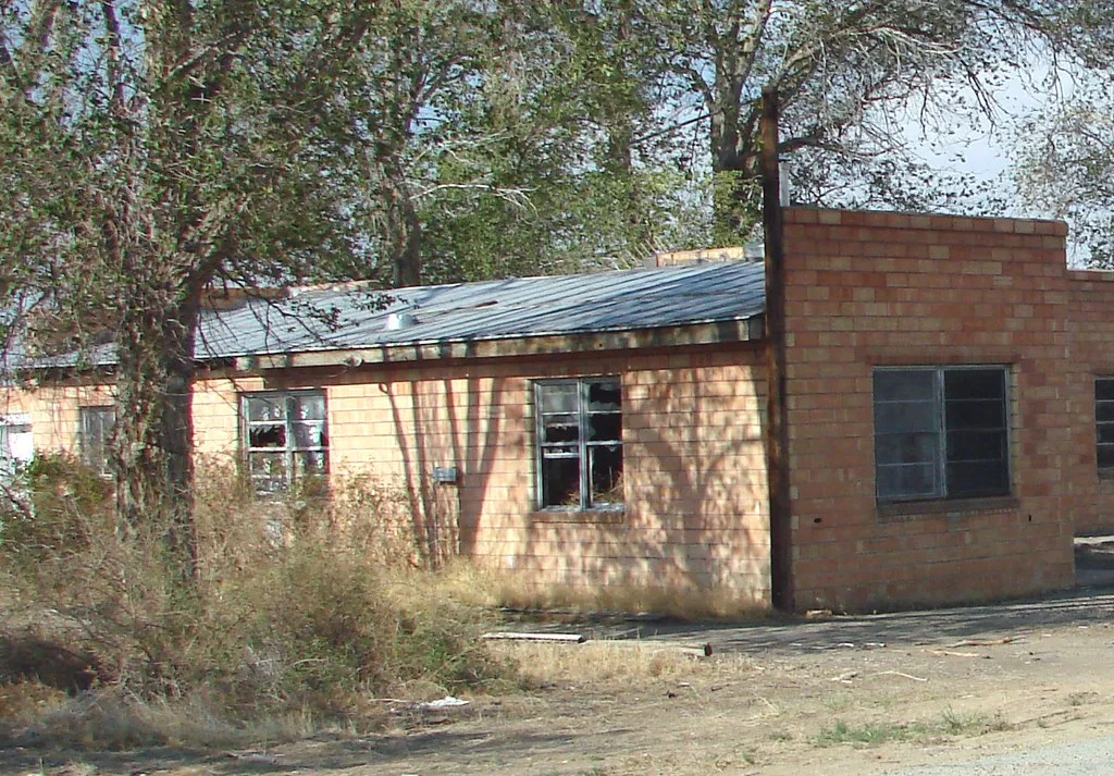 Small abandoned yellow brick building with broken windows beneath a sheet-metal roof, surrounded by trees and scrub.