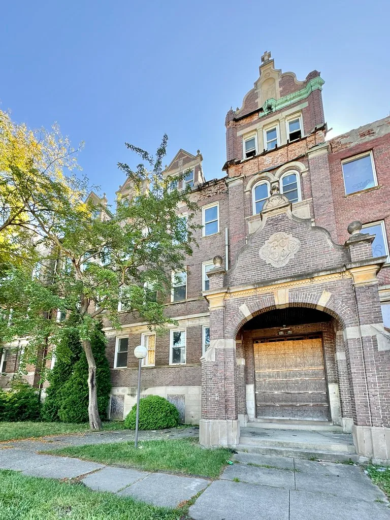 Ornate facade of an abandoned building with a barricaded entrance and deteriorated brick cornices.