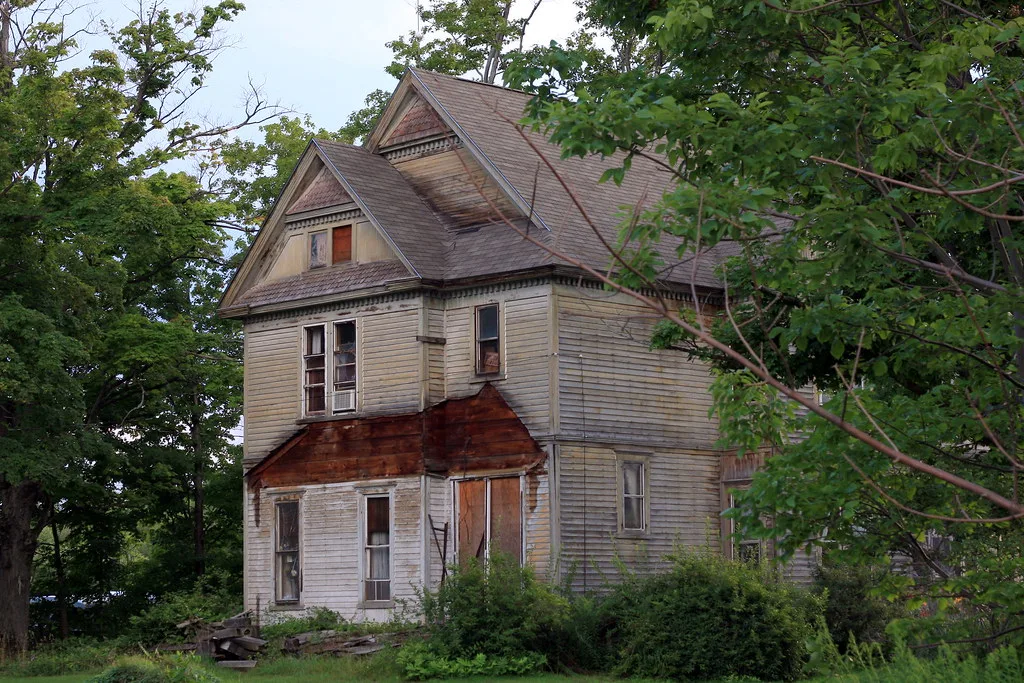Large derelict wooden house with boarded openings among bushes.