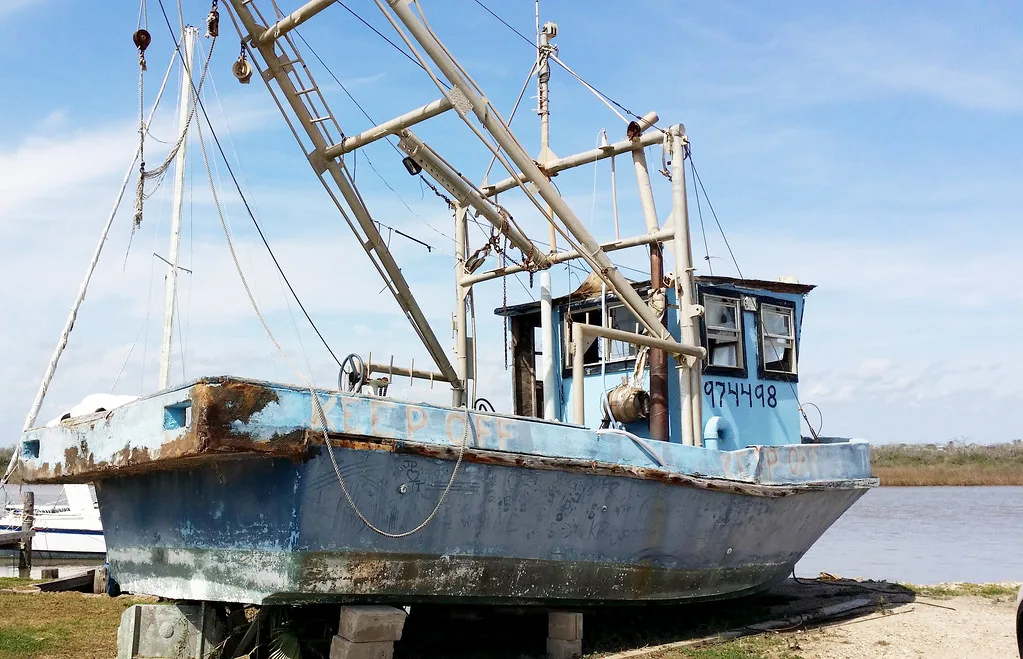 Ancien bateau de pêche bleu posé sur cales, coque rouillée et cabine aux ouvertures détériorées.