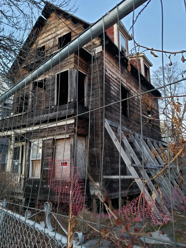 Maison en bois délabrée aux fenêtres vides, étayée derrière une clôture.