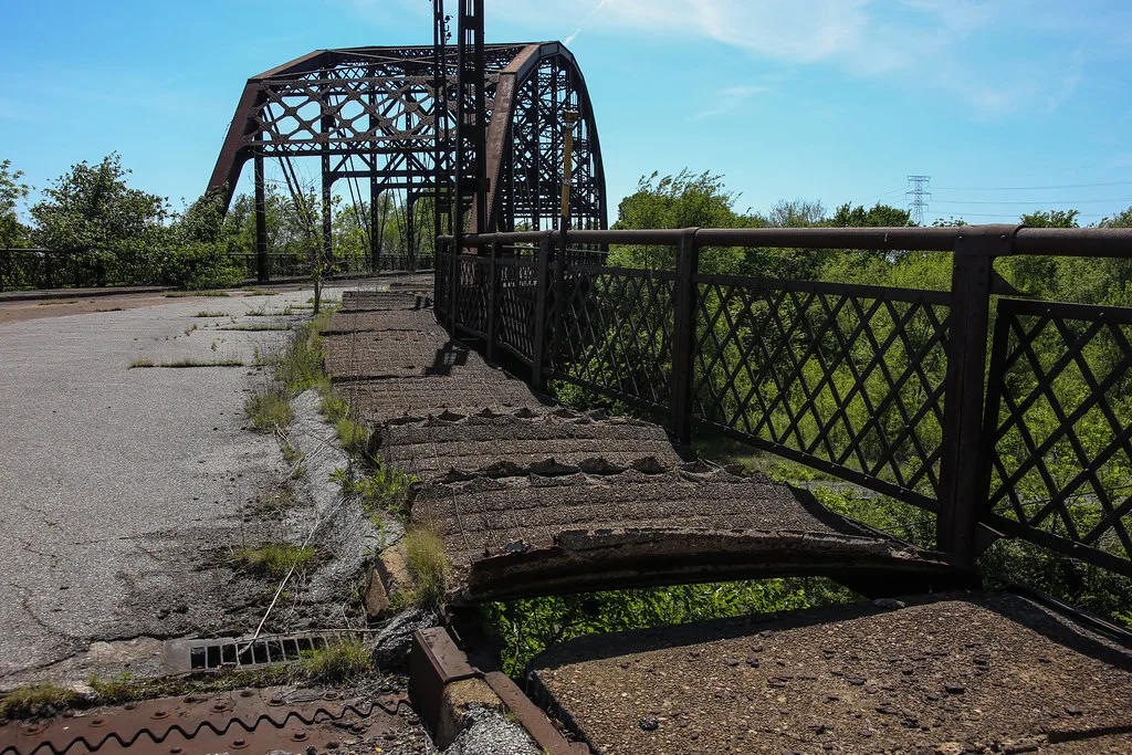 Rusty metal bridge with a broken sidewalk and weeds in the cracked roadway.