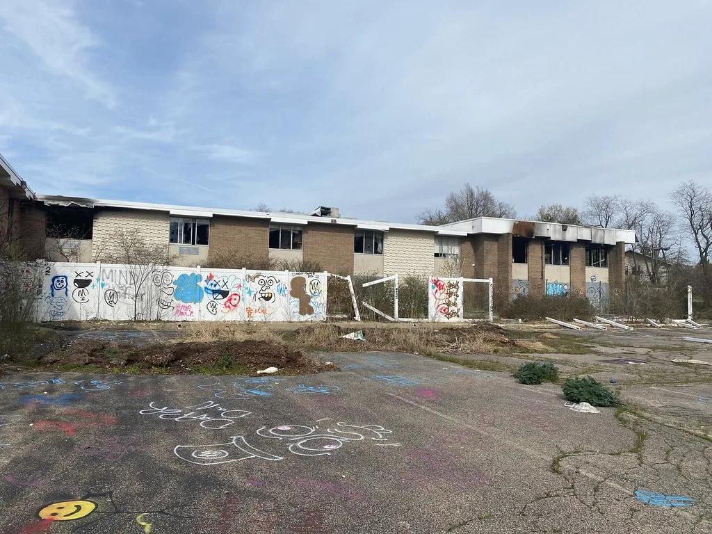 Burnt two-storey brick building behind a graffiti-covered fence and weeds.