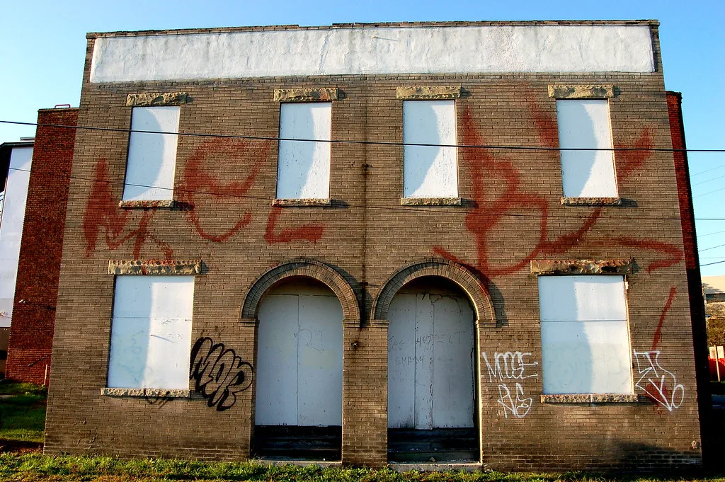 Façade de briques aux fenêtres condamnées et deux entrées en arc, couverte de tags.