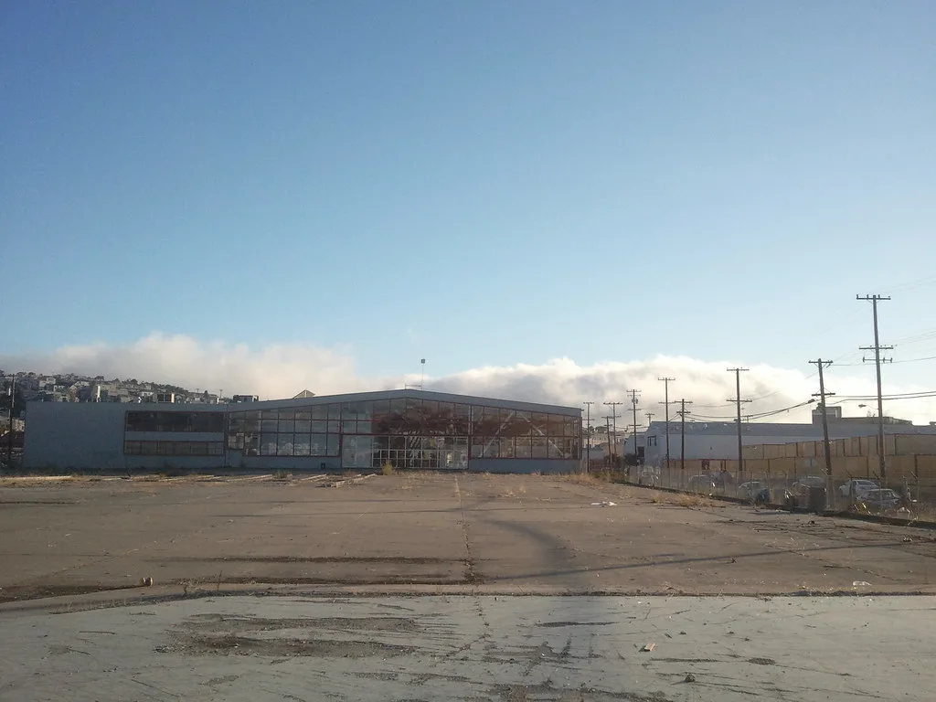 Large abandoned industrial building with broken glazing and holes in its roof facing an empty cracked yard.