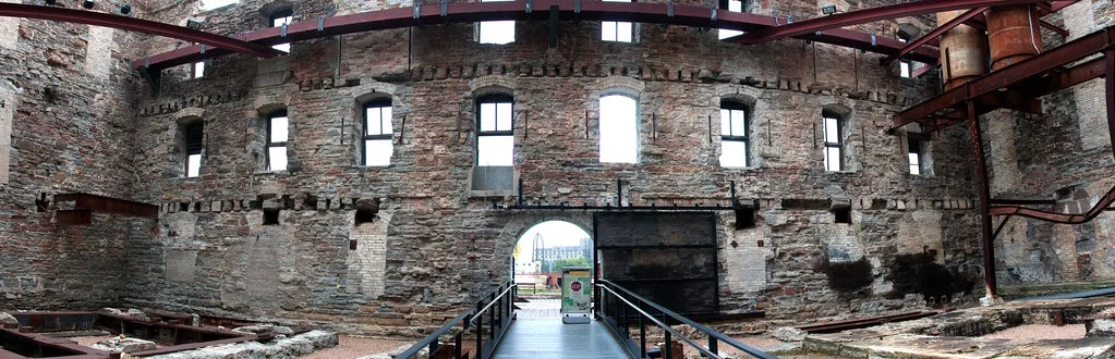 Interior of a stone industrial ruin with metal beams and a central walkway.