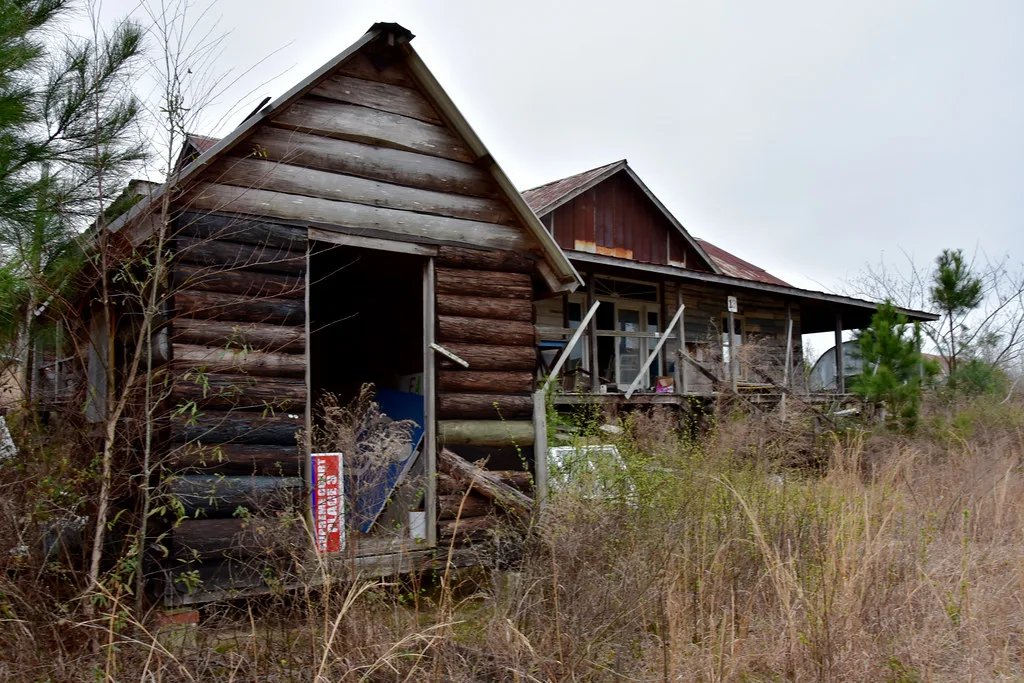 Small log cabin with an empty doorway before a wooden house overgrown with tall weeds.