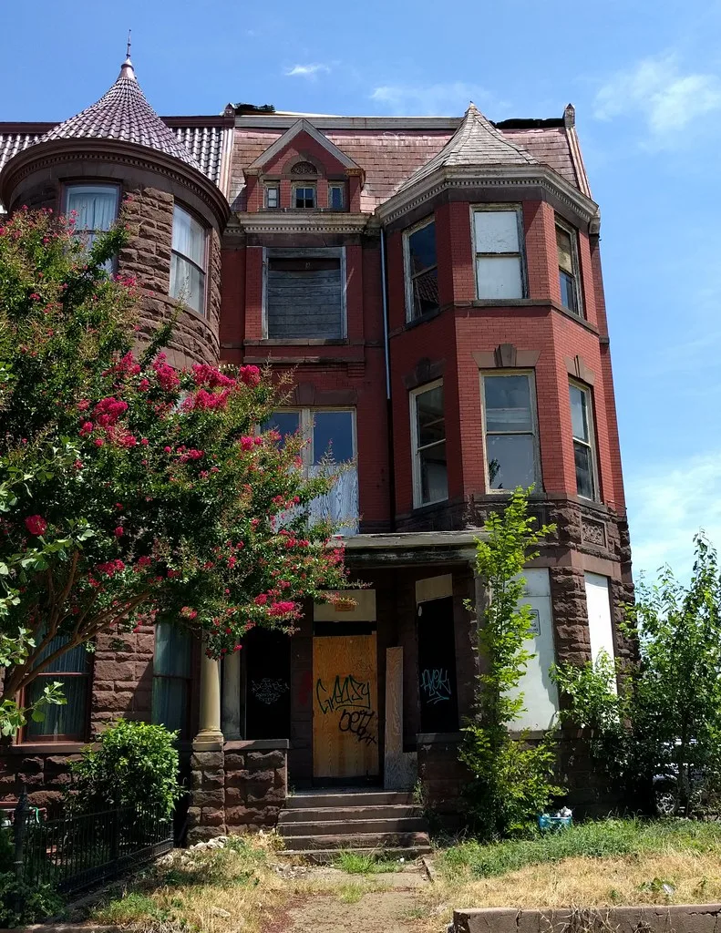 Abandoned red brick house with a turret, broken or boarded windows and a graffiti-covered entrance, surrounded by shrubs and grass.