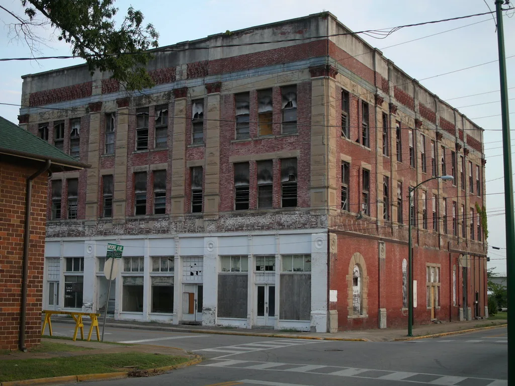 Three-storey abandoned brick building with many broken windows and boarded white shopfronts on a street corner.