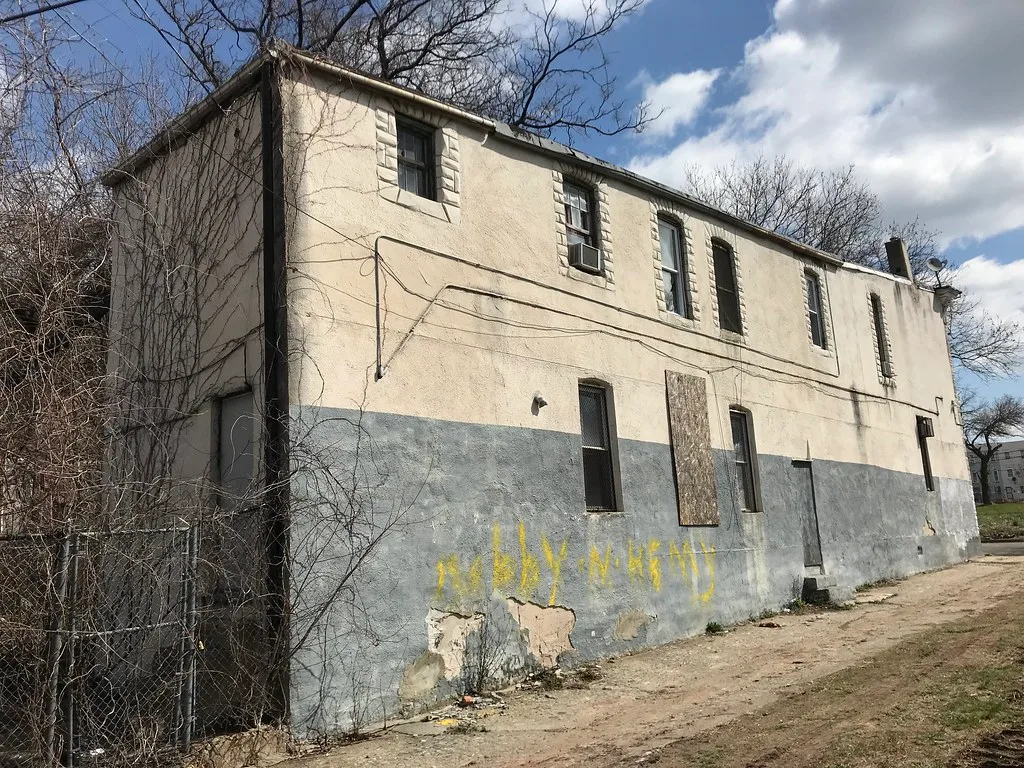 Old beige and grey building with partly boarded openings and deteriorated walls.