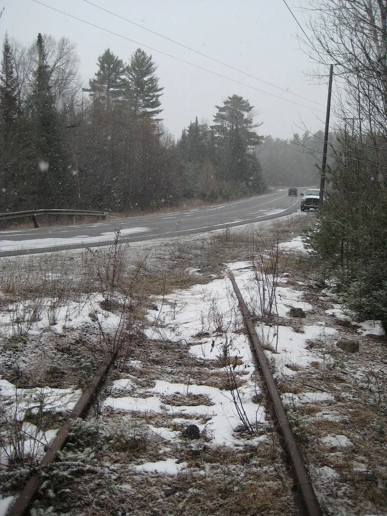Rails rouillés dans les herbes et la neige au bord d’une route.