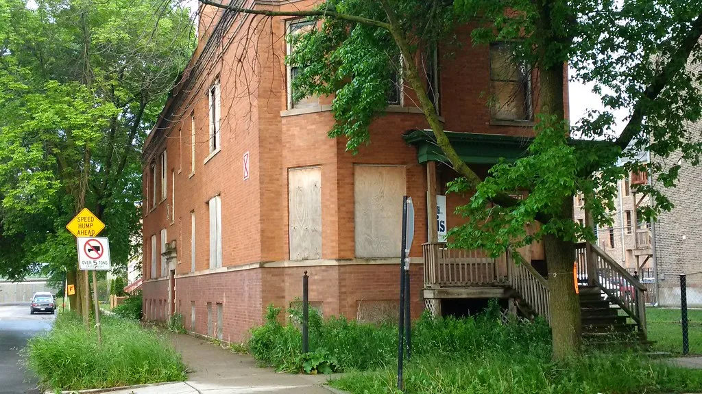 Abandoned brick house with boarded windows and a front porch overgrown by trees.