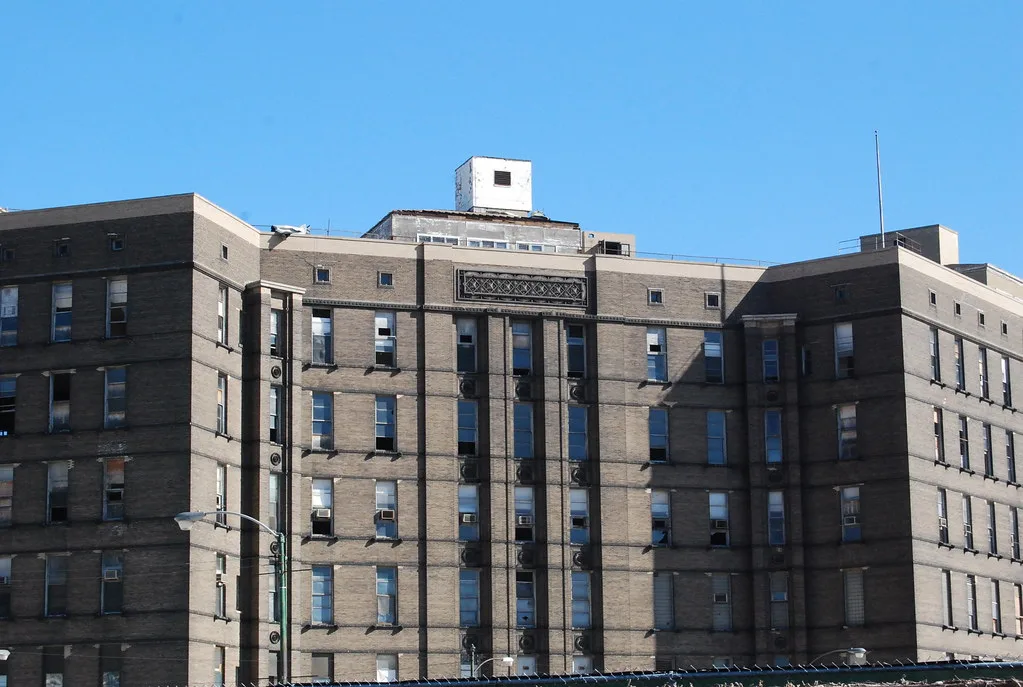Facade of a large disused brick building with several broken or sealed windows.