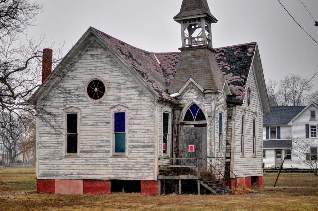 Abandoned white wooden church with an open bell tower, damaged roof and boarded door above broken steps.