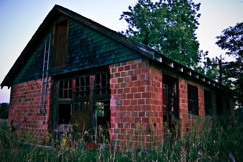 Small red brick building with broken doors and windows in tall grass.