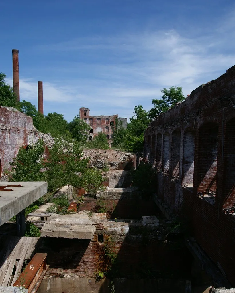 Brick factory ruins with two chimneys, empty window openings and overgrown foundations.