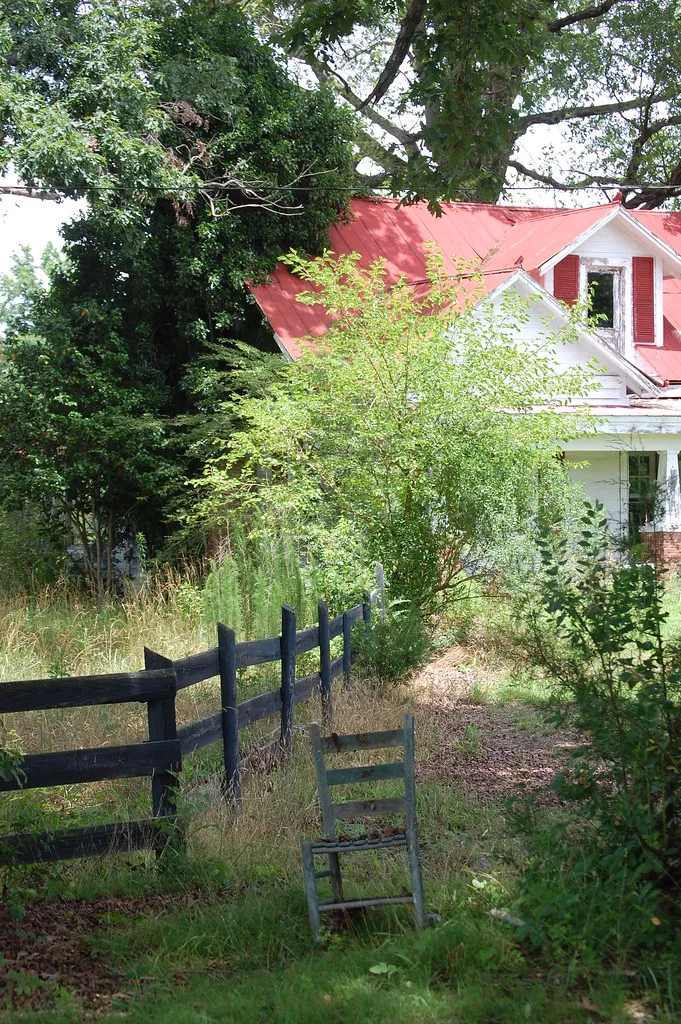 Abandoned red-roofed house partly hidden by shrubs, with a discarded chair in the grass.