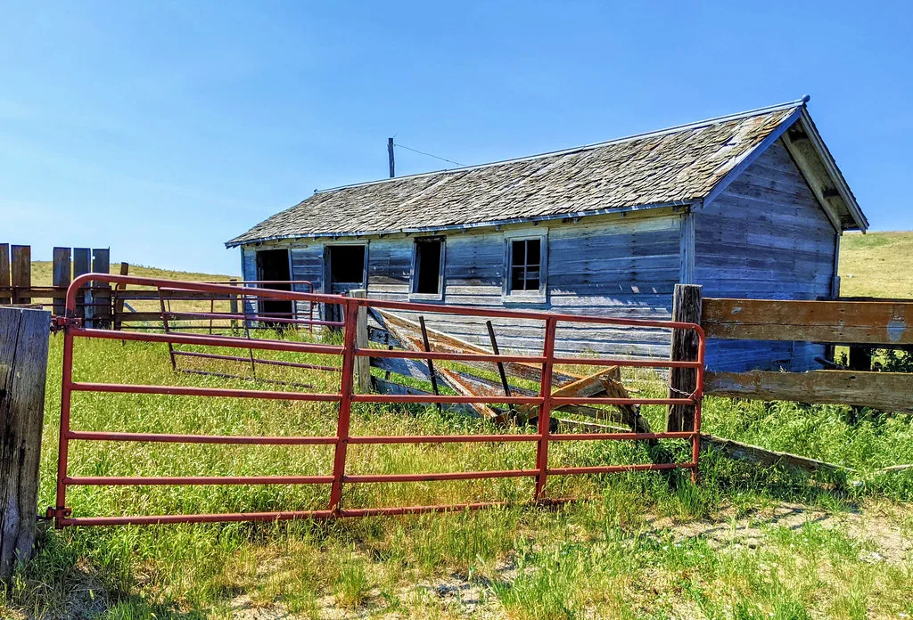 Small abandoned blue wooden building with empty windows and a deteriorated roof behind a red gate and broken fence.