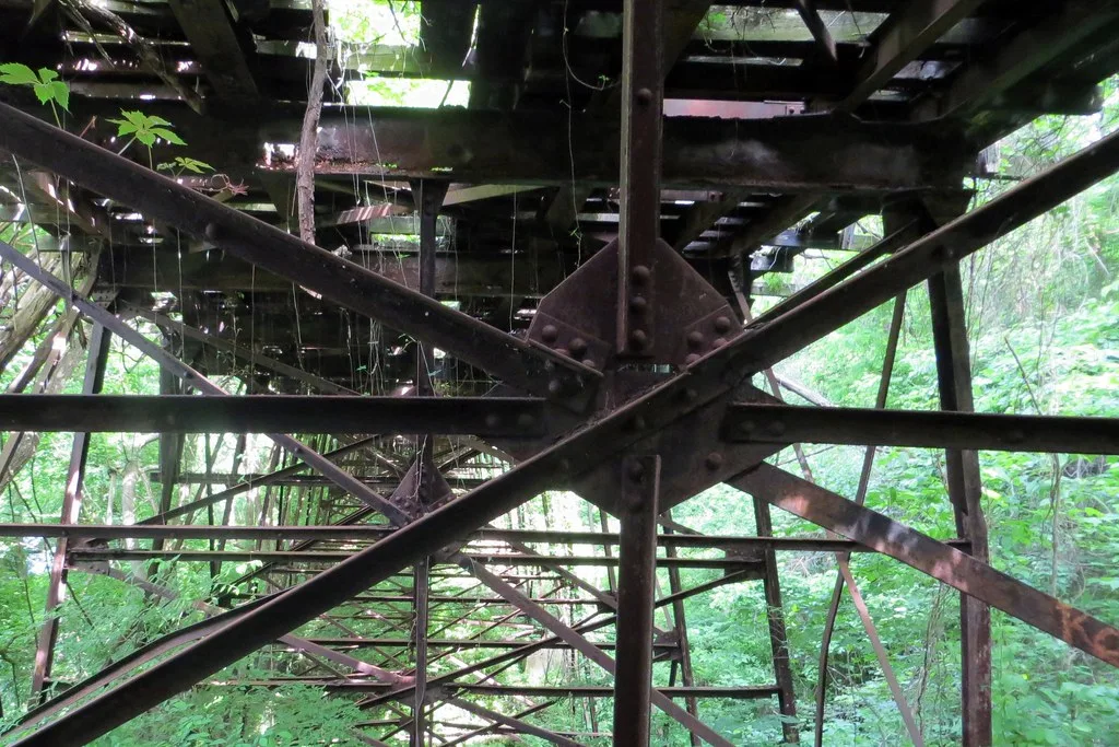 Charpente métallique rouillée sous un ancien pont envahi de végétation.