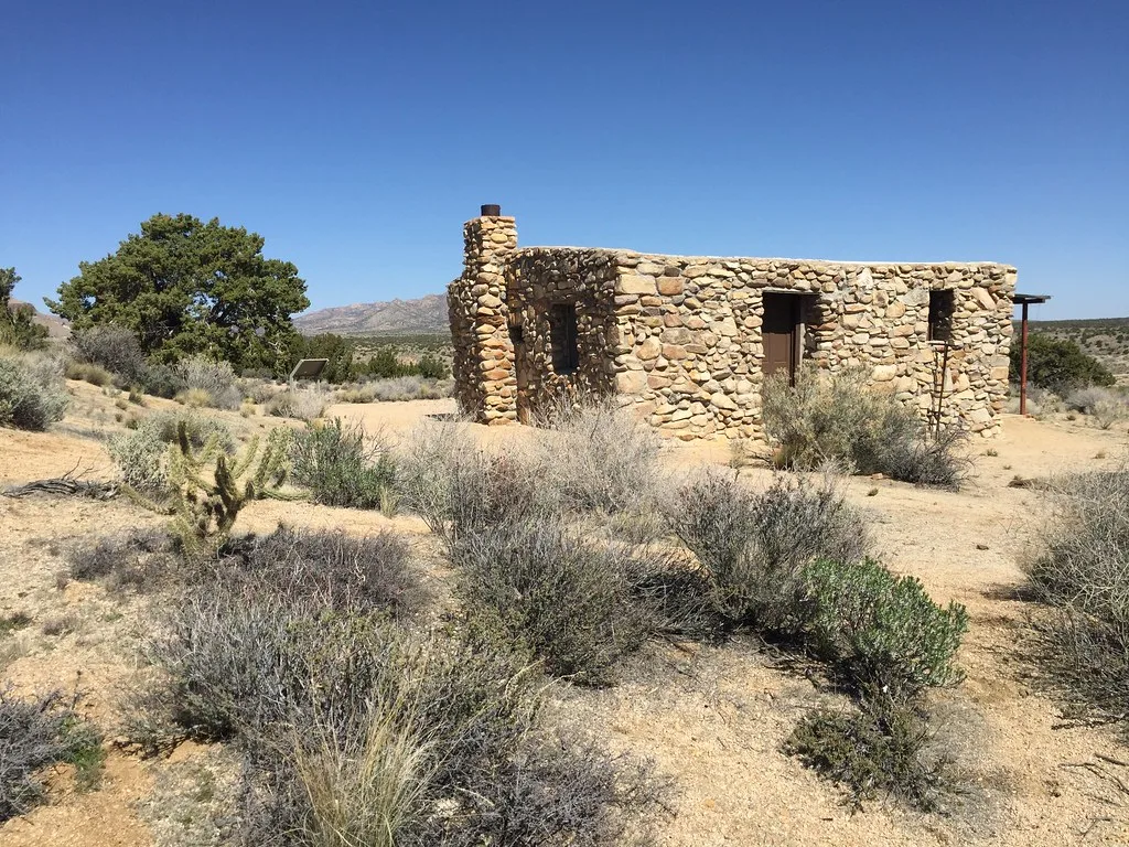 Roofless stone ruin with a remaining chimney among shrubs and cacti in an arid landscape.