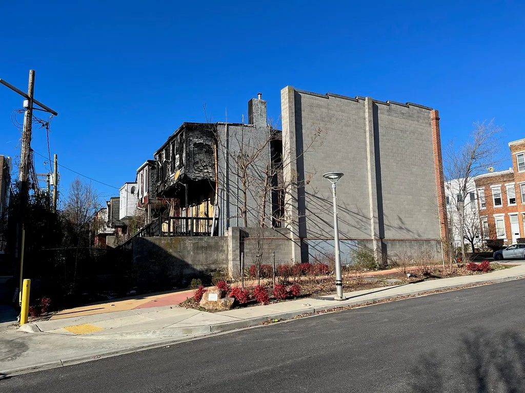 Derelict urban house with a blackened facade and empty windows beside a tall concrete-block wall.