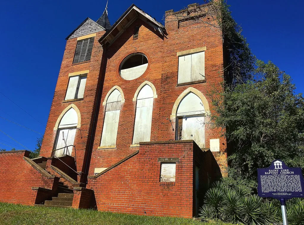 Old red-brick church with boarded openings and a tower overgrown with vegetation.