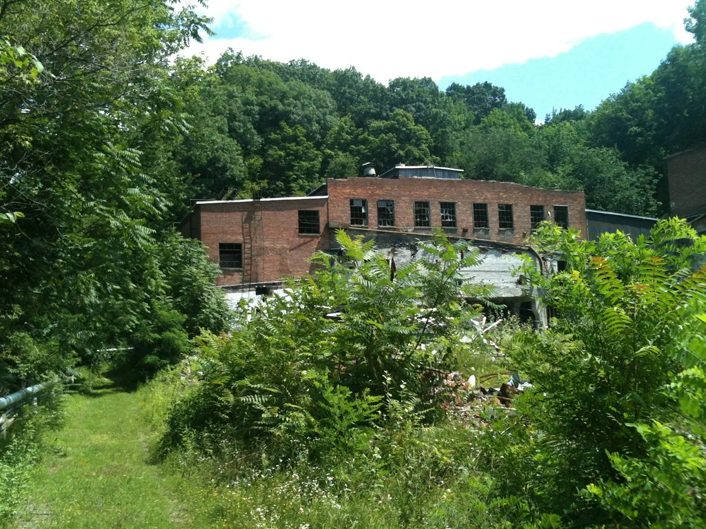 Abandoned brick factory with broken windows, behind a deteriorated annex and dense shrubs among trees.