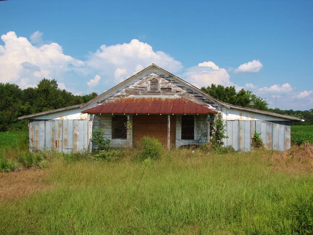 Abandoned building with a rusty porch roof and metal wings surrounded by tall grass.