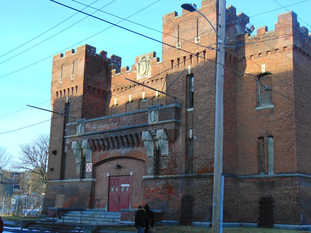 Old brick building with crenellated towers and boarded windows behind overhead cables.