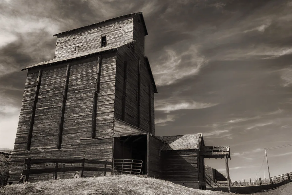 Old wooden grain elevator with weathered siding and a partially collapsed awning in a rural landscape.