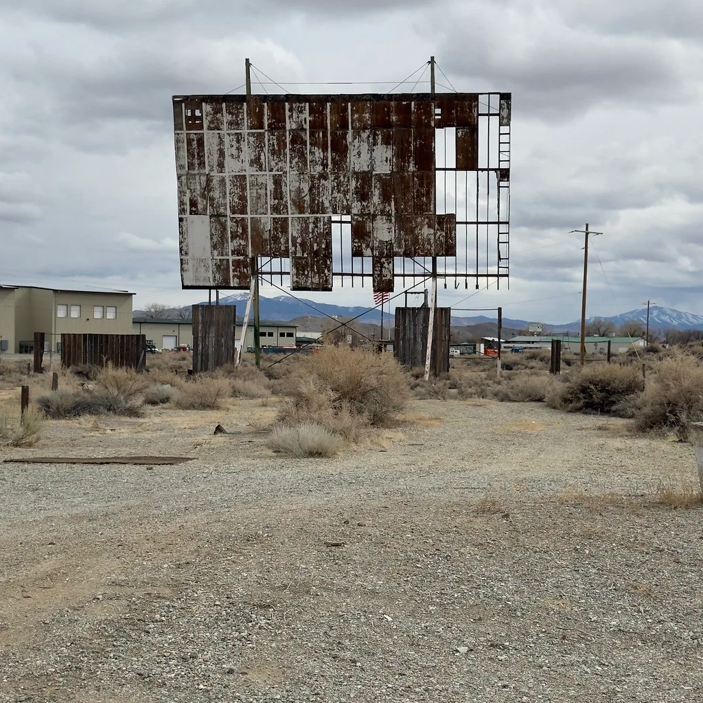 Abandoned drive-in cinema screen with missing rusty metal panels on scrub-covered ground.
