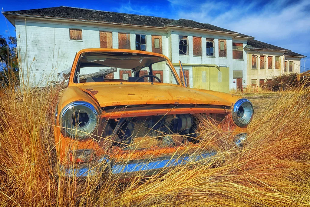 Orange vehicle wreck in grass in front of a building with boarded windows.