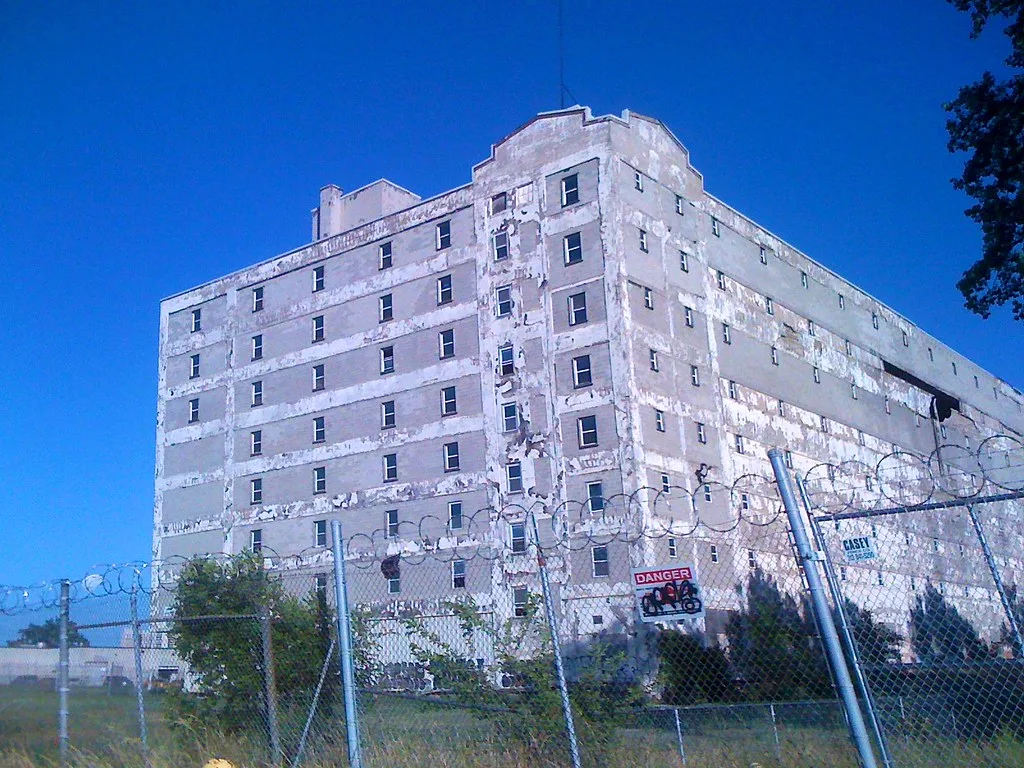 Grand bâtiment industriel au parement écaillé derrière une clôture de barbelés.