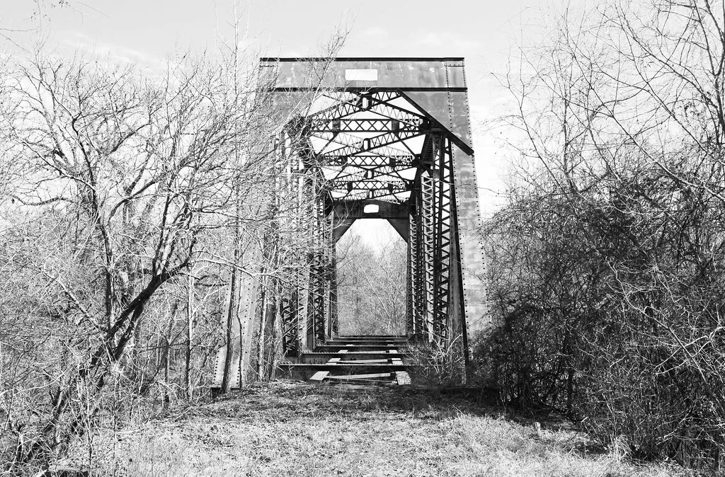 Abandoned steel bridge without a continuous deck among bare trees, in black and white.